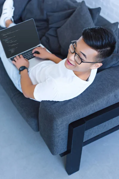 Высокоугольный вид красивого мужчины, лежащего на диване и использующего дома ноутбук с HTML-кодом — стоковое фото