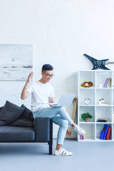Excited handsome asian man using laptop on sofa and gesturing in living room — Stock Photo