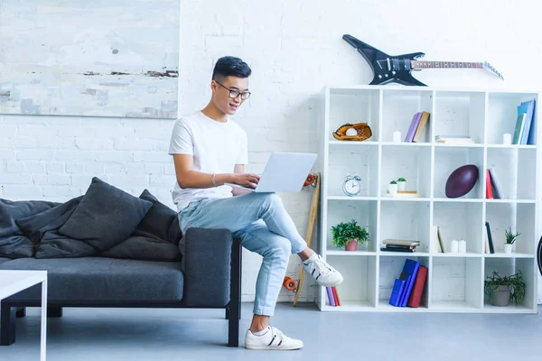 Fröhlich hübsch asiatisch mann using laptop auf sofa im wohnzimmer — Stockfoto