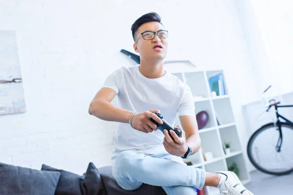 Irritated asian man playing video game with joystick on sofa at home — Stock Photo