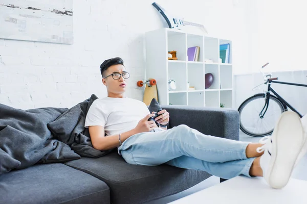 Traurig schöner asiatischer Mann spielt Videospiel auf Sofa zu Hause — Stockfoto