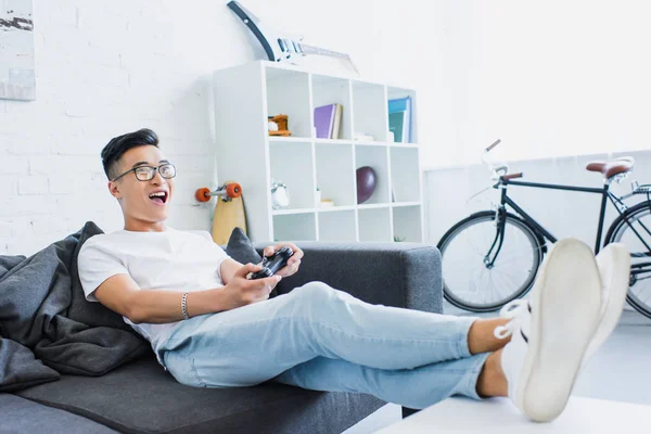 Excited handsome asian man playing video game on sofa in living room — Stock Photo