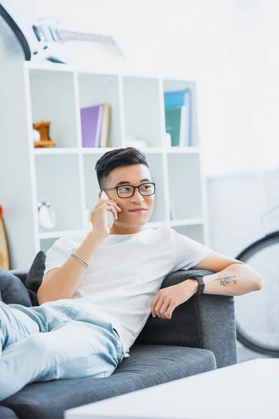 Cheerful handsome asian man lying on sofa and talking by smartphone at home — Stock Photo