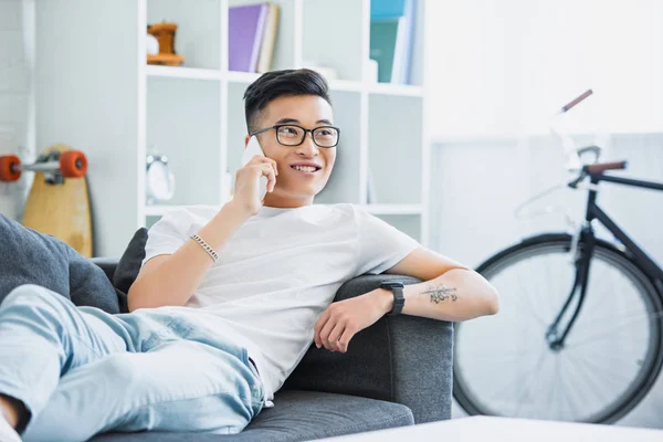 Smiling handsome asian man lying on sofa and talking by smartphone at home — Stock Photo
