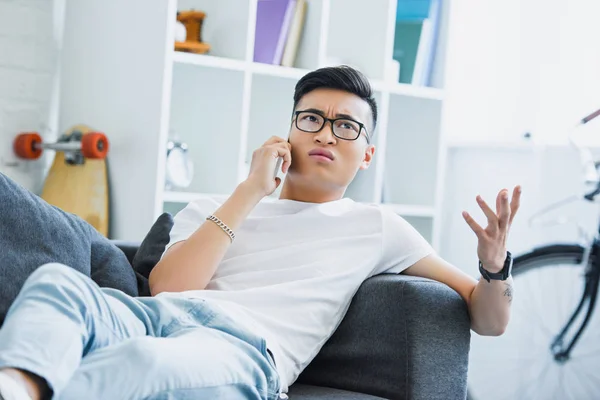 Irritado asiático hombre acostado en sofá, hablando por teléfono inteligente y gesto en casa - foto de stock