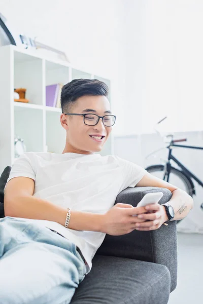 Щасливий красивий азіатський чоловік лежить на дивані і використовує смартфон вдома — стокове фото