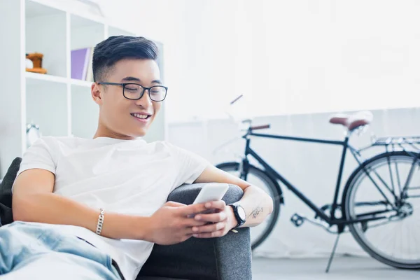 Smiling handsome asian man lying on sofa and using smartphone at home — Stock Photo
