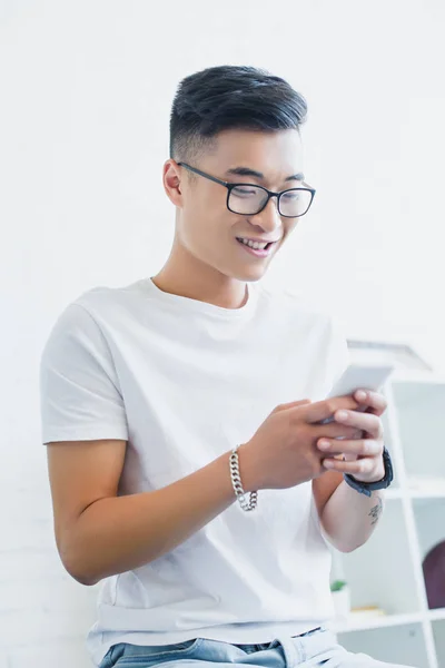 Sonriente guapo asiático hombre en gafas usando smartphone en casa - foto de stock