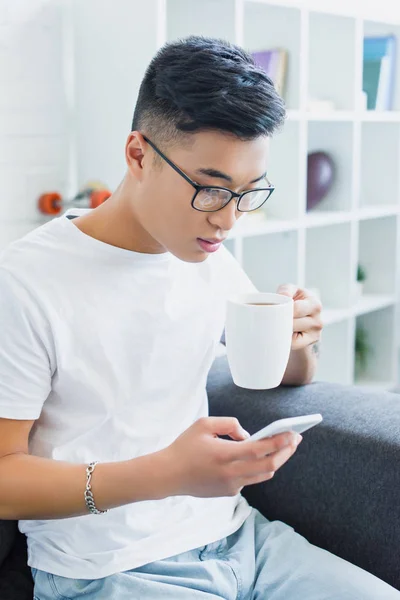 Красивый азиатский мужчина пьет чай и использует смартфон на диване дома — стоковое фото