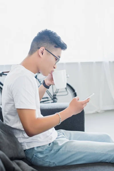 Side view of handsome asian man drinking tea and using smartphone on sofa at home — Stock Photo