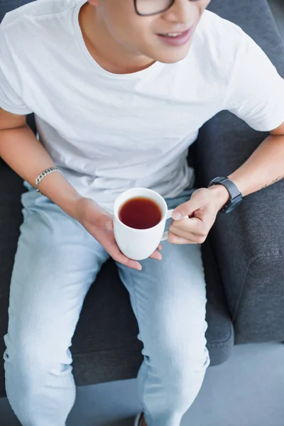 Recortado imagen de asiático hombre sentado en sofá con taza de té en casa - foto de stock