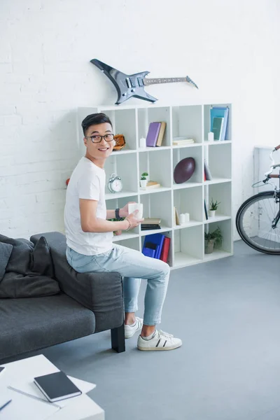 Schöner asiatischer Mann sitzt auf Sofa Seite mit einer Tasse Tee und schaut weg zu Hause — Stockfoto