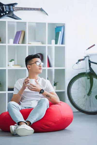 Guapo asiático hombre sentado en rojo frijol bolsa silla con taza de café y mirando lejos en casa - foto de stock