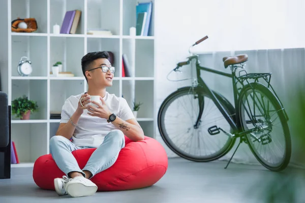 Beau asiatique homme assis sur haricot sac chaise avec tasse de thé et regarder loin à la maison — Photo de stock