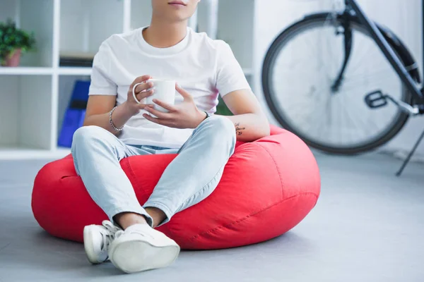 Cropped image of man sitting on red bean bag chair with cup of tea in living room — Stock Photo