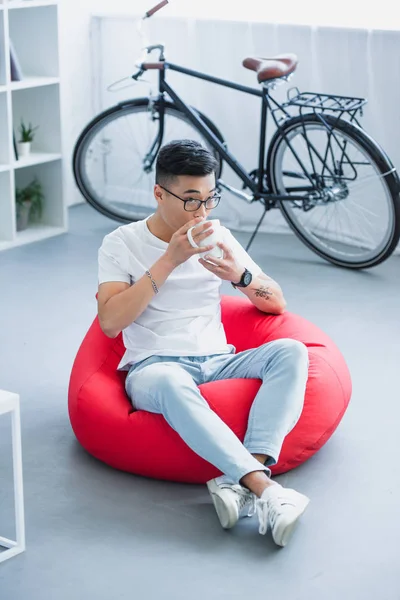 Alto ángulo vista de guapo asiático hombre sentado en frijol bolsa silla y beber té en casa - foto de stock