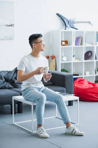 Schöner lächelnder asiatischer Mann sitzt morgens zu Hause mit einer Tasse Kaffee auf dem Tisch — Stockfoto