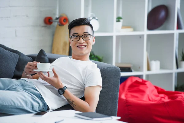 Handsome happy young asian man in eyeglasses holding cup of coffee while lying on couch — Stock Photo