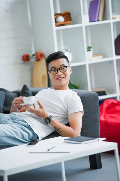 Bello sorridente giovane asiatico uomo in occhiali holding tazza di caffè mentre sdraiato su divano — Foto stock