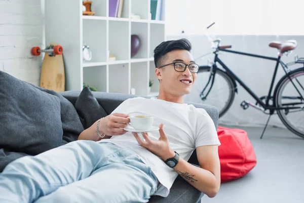 Улыбающийся молодой азиат в очках, держа чашку кофе и отводя взгляд, лежа на диване — стоковое фото