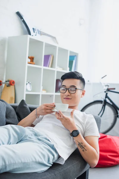 Happy young asian man in eyeglasses lying on couch and holding cup of coffee — Stock Photo