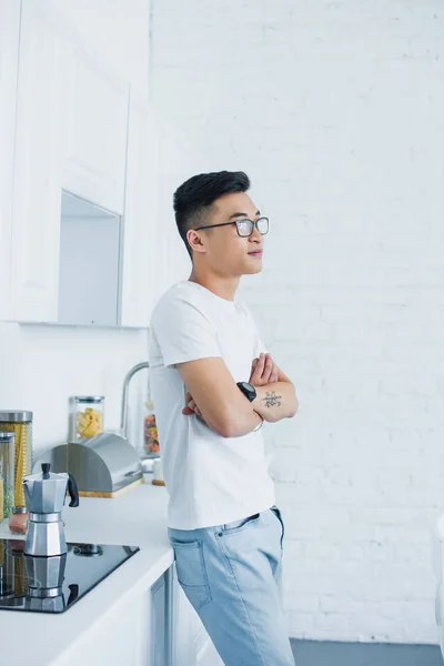 Vue latérale de beau jeune homme asiatique dans des lunettes debout avec les bras croisés et regardant loin dans la cuisine — Photo de stock