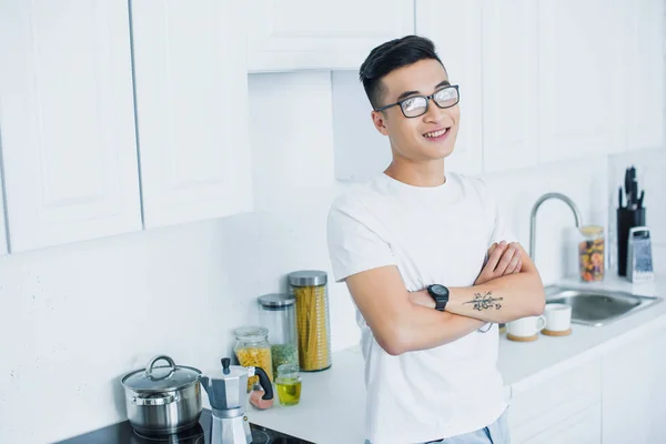 Bello sorridente giovane asiatico uomo in occhiali in piedi con le braccia incrociate in cucina — Foto stock