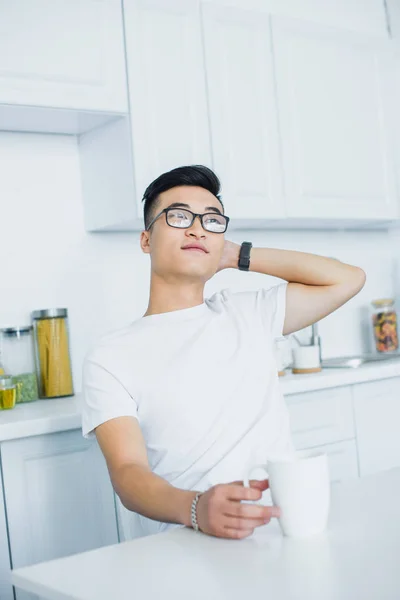 Schöner junger asiatischer Mann mit Brille, der eine Tasse Tee hält und wegschaut, während er in der Küche sitzt — Stockfoto
