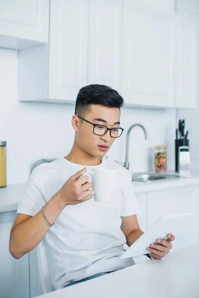 Young asian man in eyeglasses holding cup and using digital tablet — Stock Photo