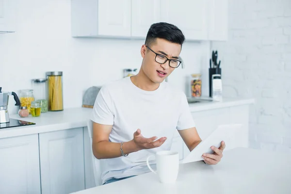 Emotional young asian man in eyeglasses using digital tablet while sitting in kitchen — Stock Photo