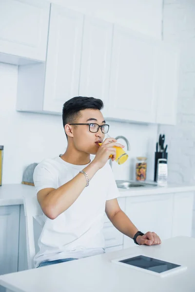 Giovane asiatico uomo in occhiali bere succo e guardando lontano in cucina — Foto stock