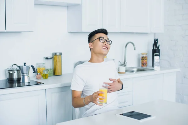 Feliz jovem asiático homem segurando vidro de suco e olhando para cima enquanto sentado na cozinha — Fotografia de Stock