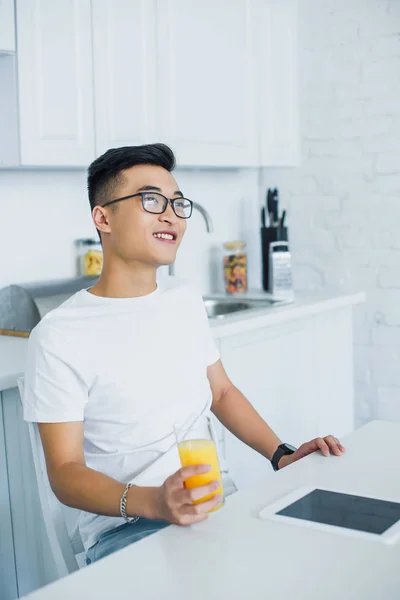 Guapo sonriente joven asiático hombre en gafas celebración de vidrio fo jugo y mirando hacia otro lado mientras sentado en cocina - foto de stock