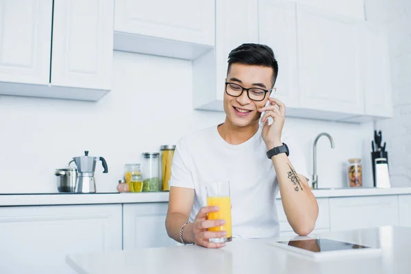 Lächelnder junger asiatischer Mann mit Brille hält ein Glas Saft in der Hand und spricht mit dem Smartphone, während er in der Küche sitzt — Stockfoto