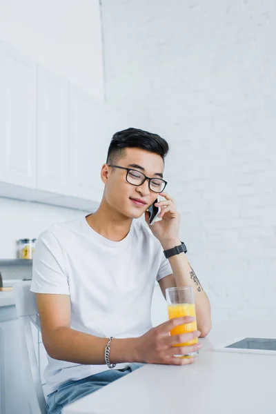 Young asian man in eyeglasses holding glass of juice and talking by smartphone while sitting in kitchen — Stock Photo