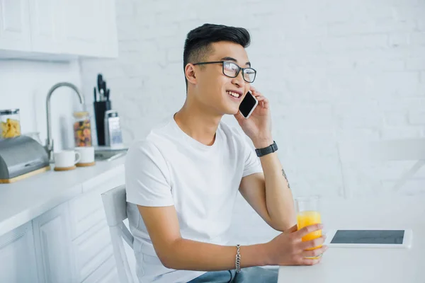 Souriant jeune homme tenant un verre de jus et parlant par smartphone tout en étant assis dans la cuisine — Photo de stock