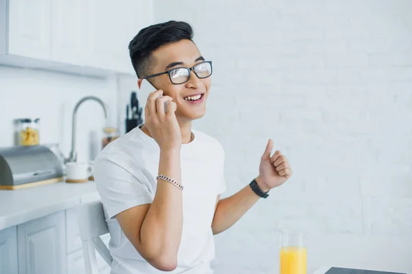 Щасливий молодий азіатський чоловік розмовляє по смартфону, сидячи на кухні — стокове фото