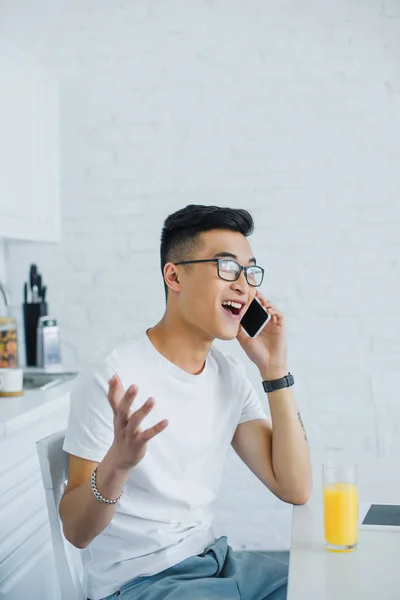 Emotional young asian man talking by smartphone while sitting in kitchen — Stock Photo