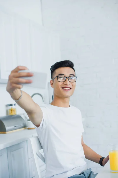 Sorrindo jovem asiático homem no óculos tomando selfie com smartphone na cozinha — Fotografia de Stock