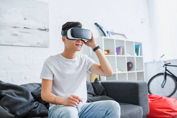 Sonriente joven asiático hombre usando virtual realidad auricular mientras sentado en sofá en casa - foto de stock