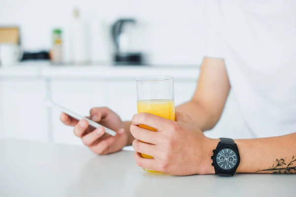 Vista parcial de cerca del joven sosteniendo un vaso de jugo y usando un teléfono inteligente - foto de stock