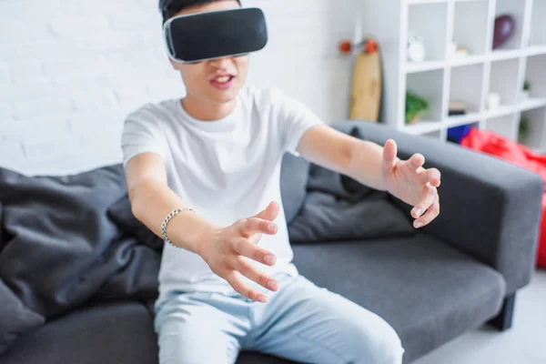 Joven asiático hombre sentado en sofá y usando virtual realidad auricular en casa - foto de stock
