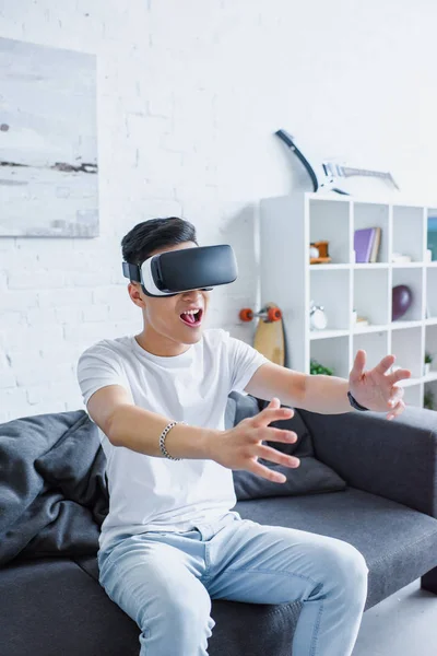 Молодой человек сидит на диване и пользуется гарнитурой виртуальной реальности дома — стоковое фото