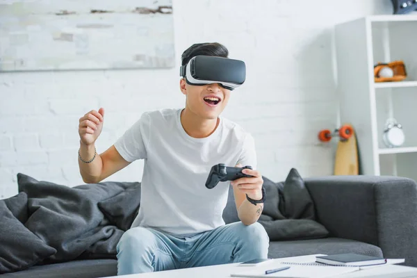 Joven feliz en auriculares de realidad virtual jugando con joystick en casa - foto de stock