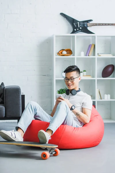 Sonriente joven asiático hombre sentado en frijol bolsa silla y usando smartphone en casa - foto de stock