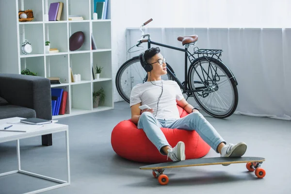 Joven asiático hombre en auriculares sentado en frijol bolsa silla y el uso de teléfono inteligente en casa - foto de stock