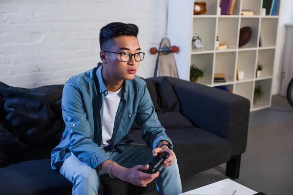 Enfocado joven asiático hombre jugando video juego con joystick en casa - foto de stock