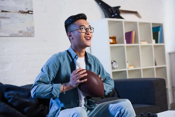 Feliz jovem asiático homem em óculos segurando bola de rugby e assistindo esporte jogo em casa — Fotografia de Stock
