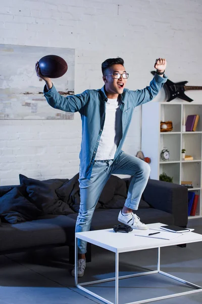 Triunfante jovem asiático homem segurando rugby bola e assistindo esporte jogo em casa — Fotografia de Stock