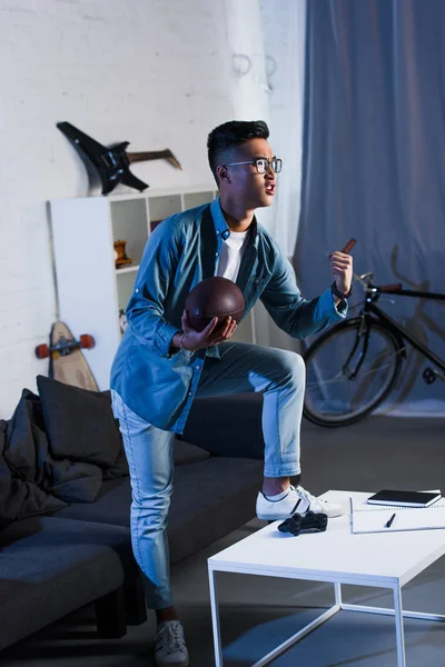 Emotionaler junger asiatischer Mann hält Rugbyball und schaut sich zu Hause ein Sportspiel an — Stockfoto